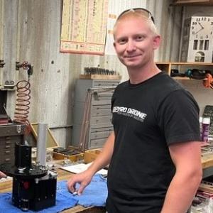 blonde man in black t-shirt stands by machined part